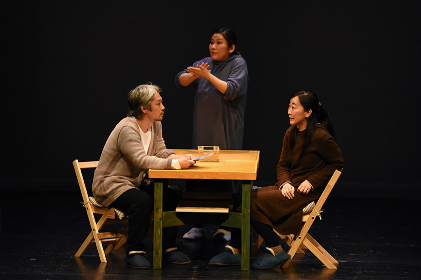 演劇『メゾン』の1シーンです。テーブルを挟んで向き合って座っている男女の俳優の間で、青い服を着た女性が手話通訳を行っています