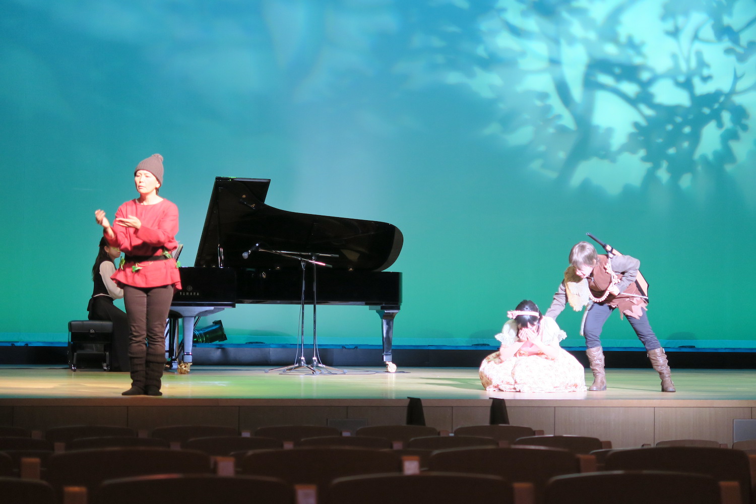 演劇『白雪姫』にて、白雪姫役と猟師役の俳優が演劇を行っている写真です。舞台の左端、ピアノの前の方で、赤い服を着た人が手話通訳を行っています