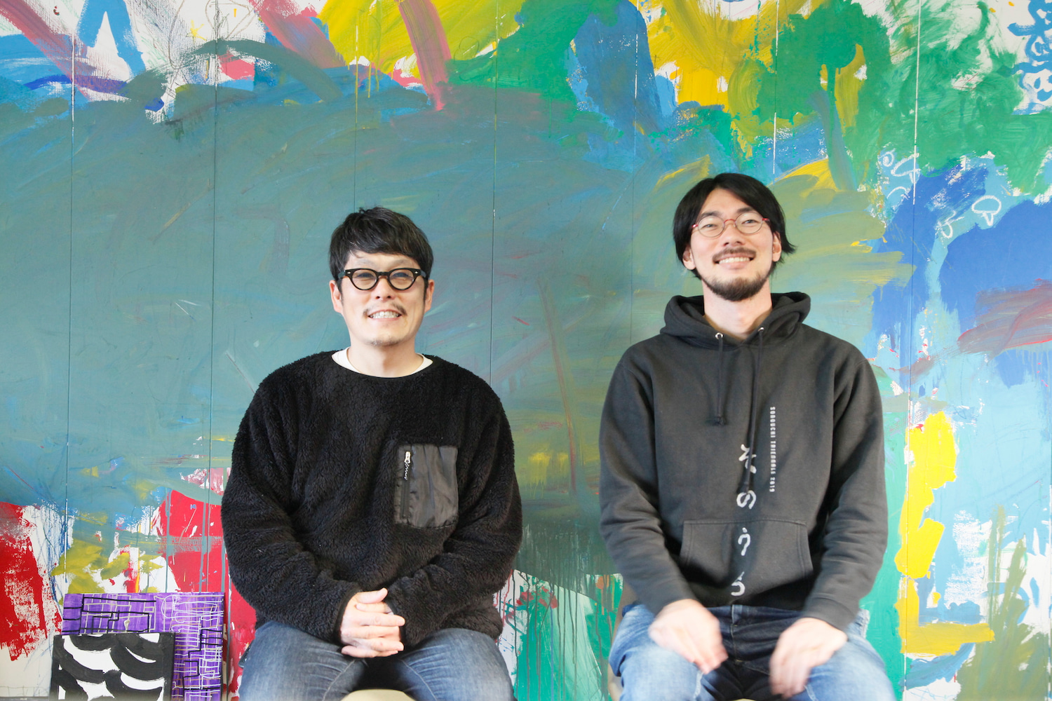 左：中野さんと右：丹正さんが、並んで座っている写真です。絵の具がカラフルに塗られた壁の前で、めがねをかけた二人が笑っています