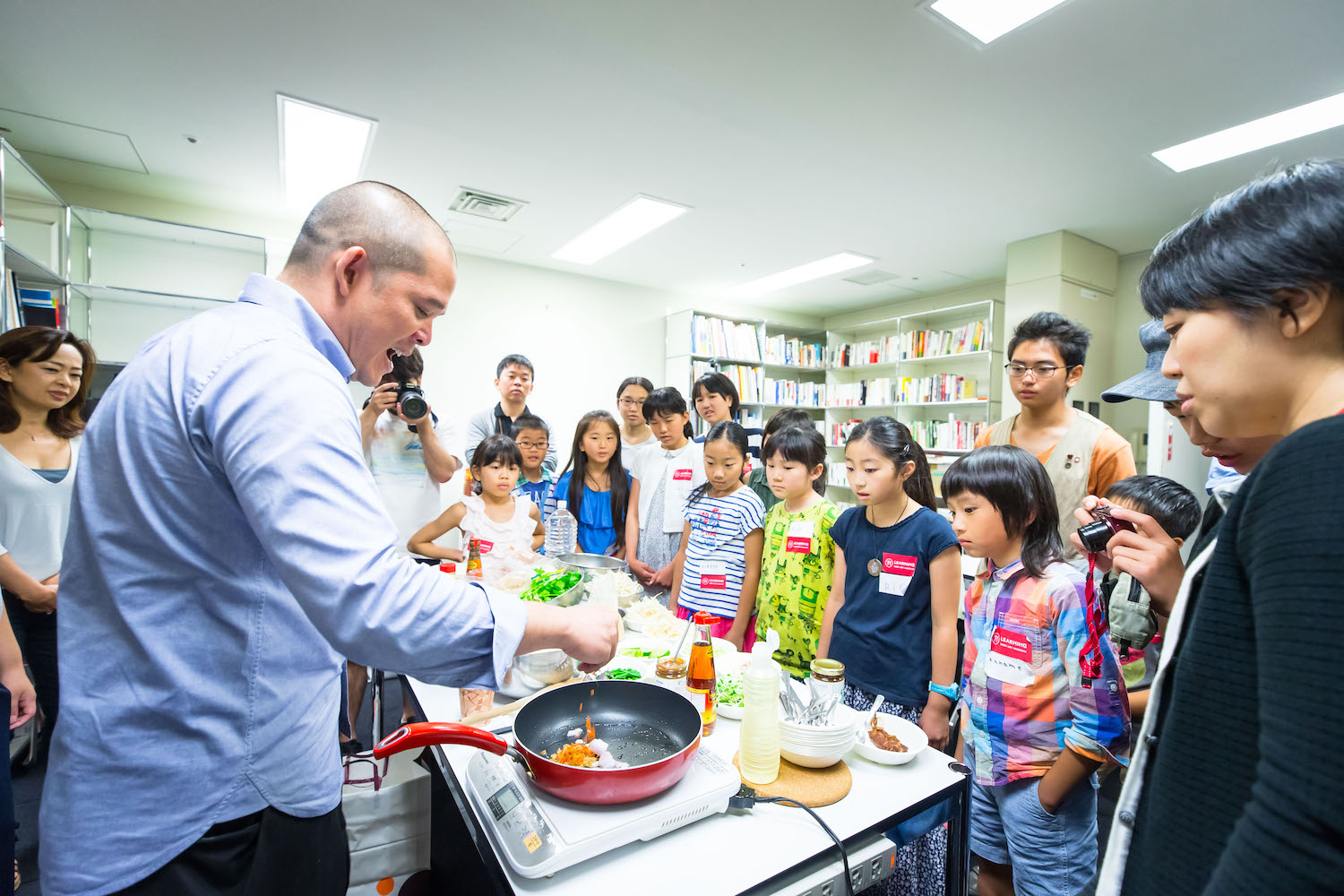 「サンシャワー展」の際に開催されたキッズ・プログラム「いろんな世代の人と一緒にアーティストに出会う」の様子です。タイのアーティスト、ドゥサディー・ハンタクーンさんがフライパンで料理をしている様子を子供たちが見つめています