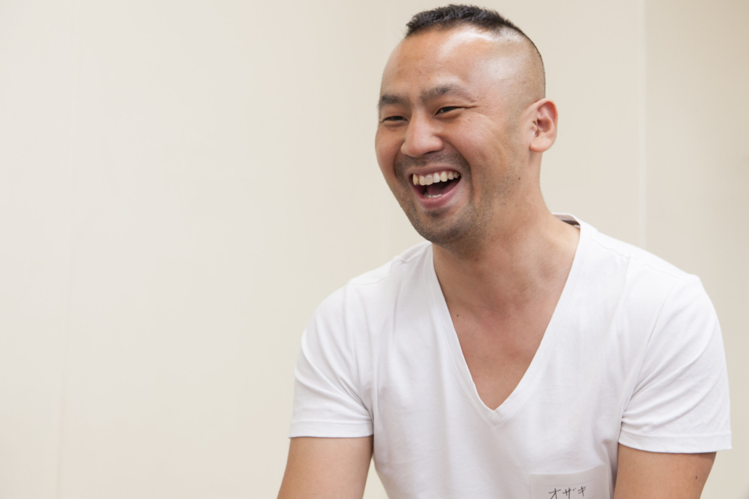 尾崎さんが白いVネックTシャツを着て口を開けて笑っている写真です。尾崎さんは頂部が短髪で、横と後ろを剃り上げたGIカットがトレードマークです