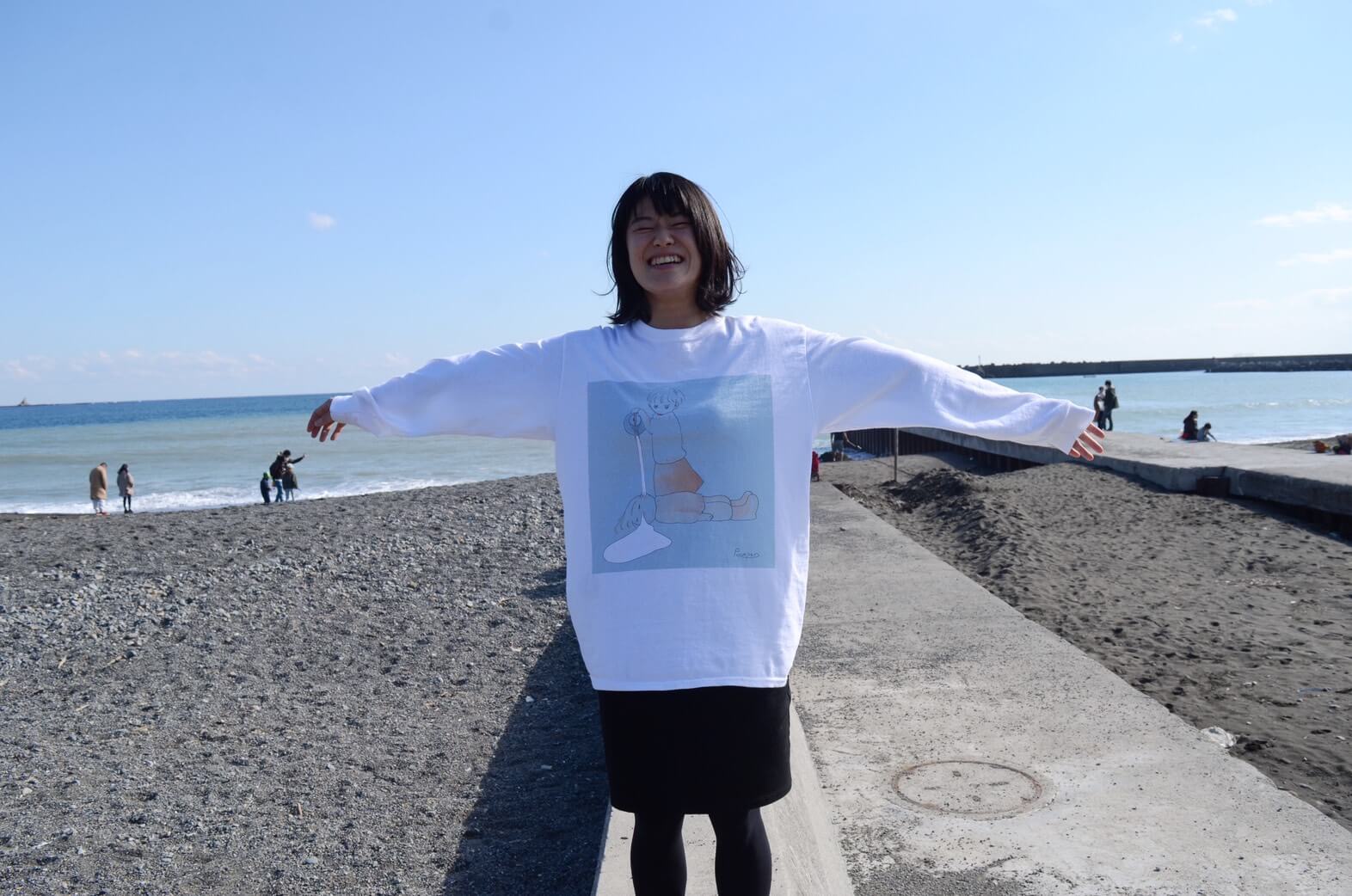 豊川弘恵さんのポートレート写真。白いシャツに黒いスカートをはいた女性が、両手を広げ海岸を背景にポーズをとっている