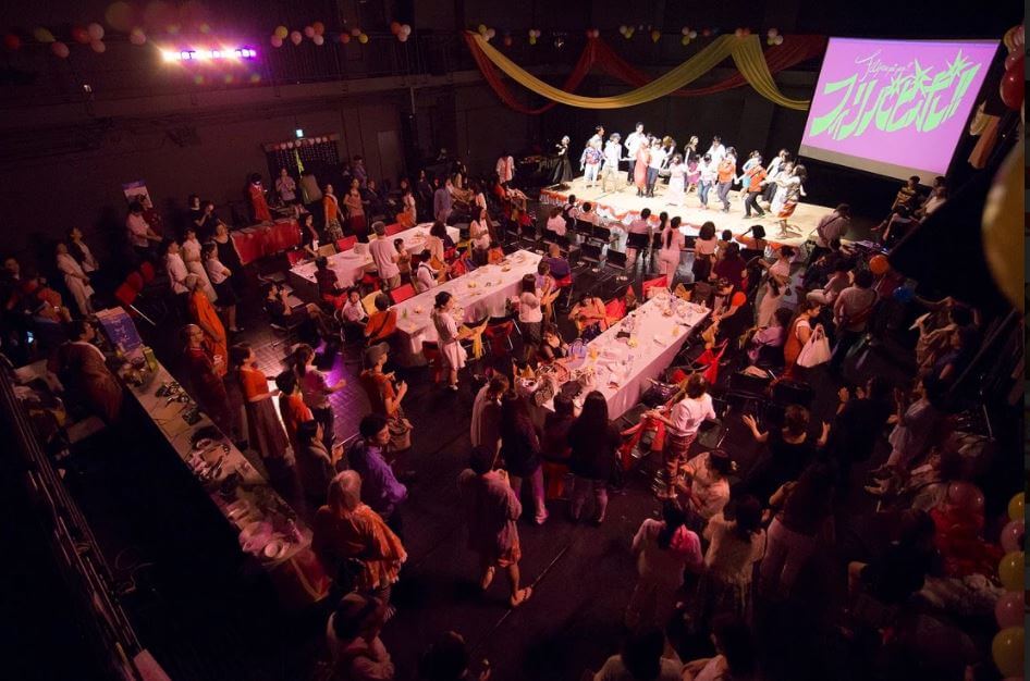「フィリパピポ!!」の東京藝術大学の千住キャンパスのホールでの様子を上から捉えた写真。日本やフィリピンのみならず、さまざまな国籍の参加者・出演者たち200名以上が参加。舞台の前に観客席が2列あり、その後ろは12人掛けの長方形のテーブルが3つ並んでいる。テーブルにはテーブルクロスがかかり、上に飲み物などがある。着席している人は一部で、ほとんどの人が立って、舞台を鑑賞している