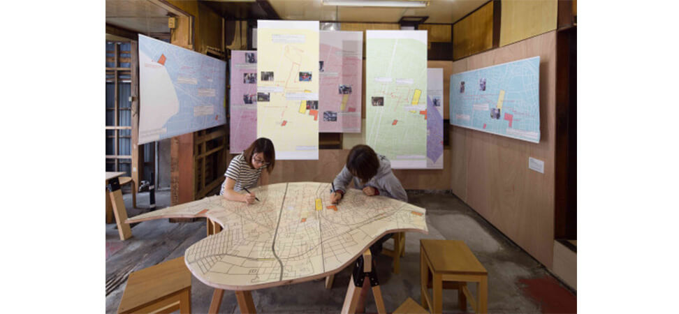 会場１階の様子。千住の地形を型取った大きな流線型のフォルムの木造りのテーブルの表面に地図が描かれており、二人が机へ向かって作業をしている
