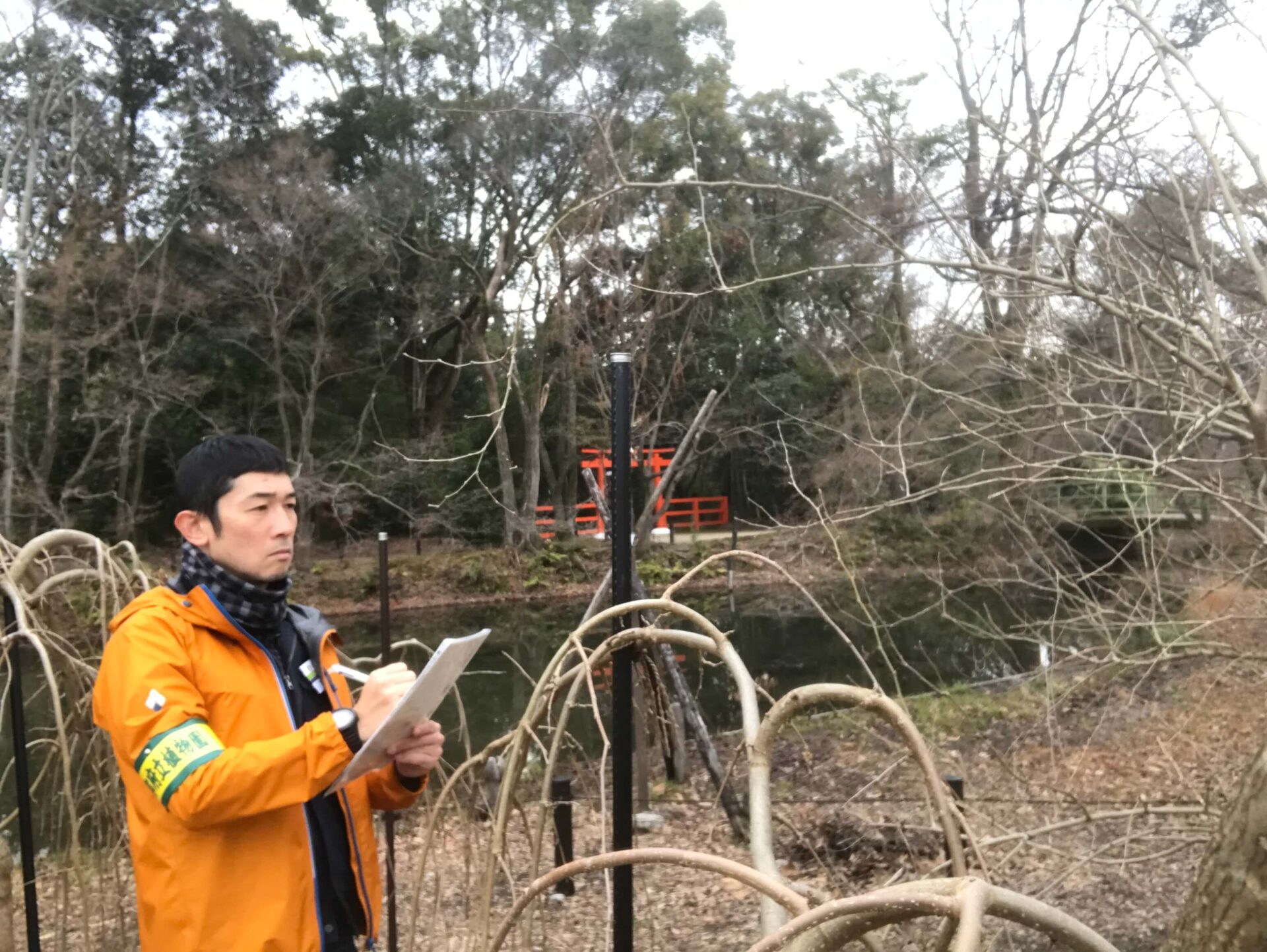 竹浪さんが、植物園で調査をされた際のお写真です。オレンジ色のジャンパーを着た竹浪さんが、植物園の木々の前で、手に持ったノートに何か書き込みをしています