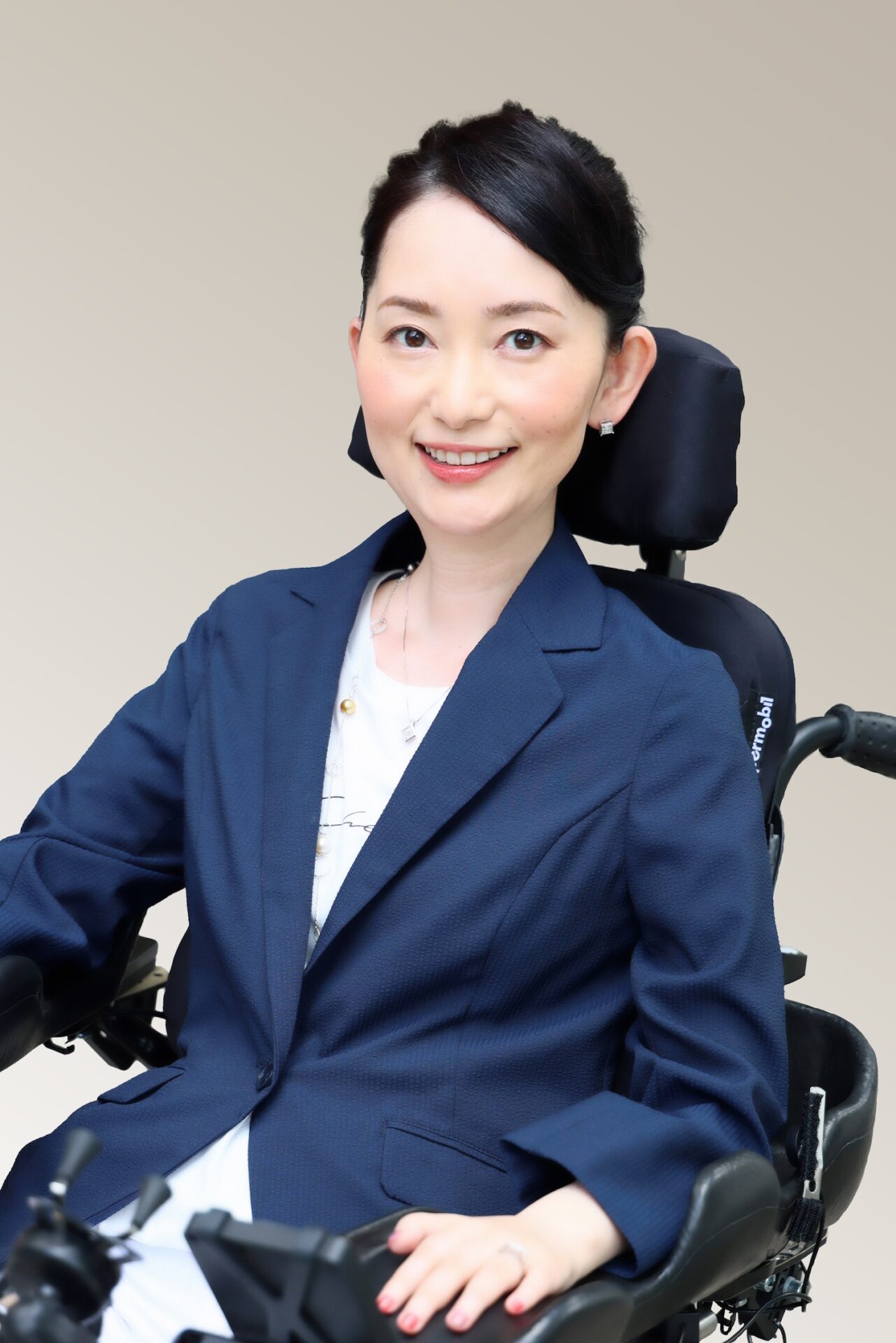 車椅子に乗った小野さんが、笑顔でこちらを見つめています。黒髪を後ろにまとめ、紺のジャケットを着ています