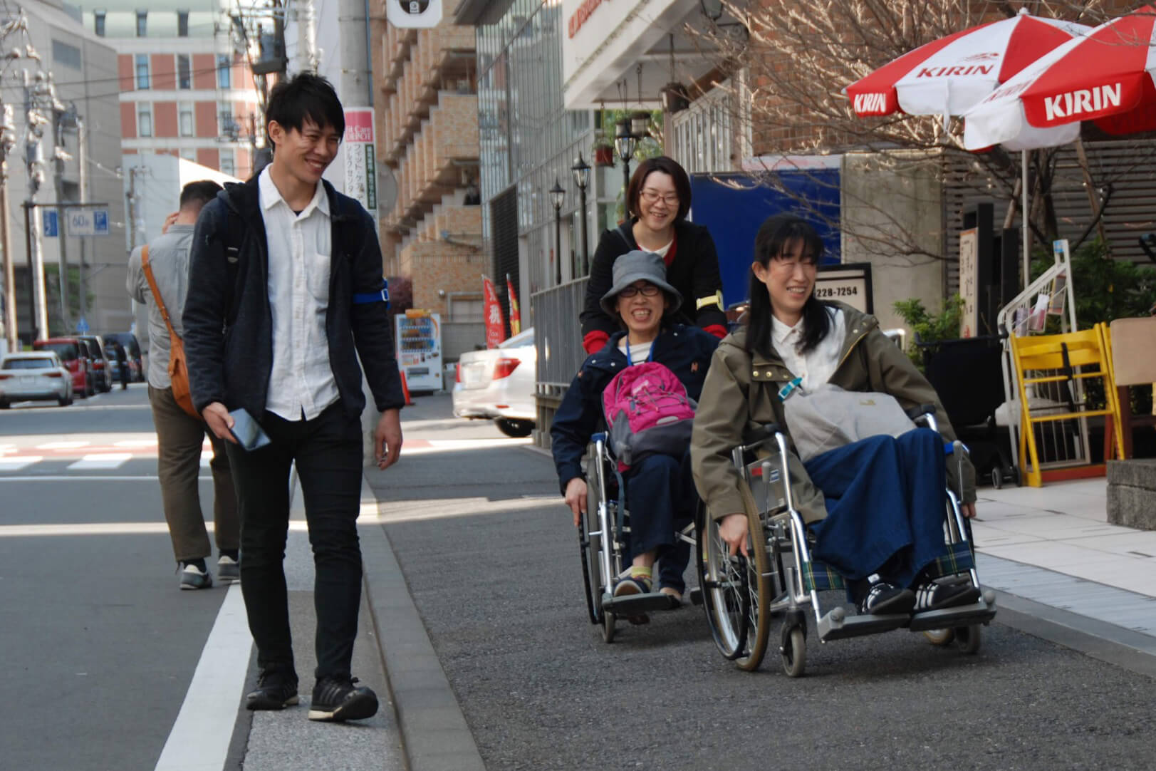 車椅子体験をしている様子を写した写真です。車椅子に乗った2名の女性と車椅子を押す1名の女性、車椅子の横を歩く1名の男性が笑顔でまちの歩道を進んでいます