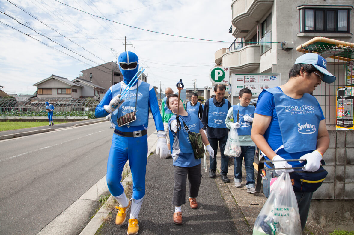 ゴミコロレンジャーと参加者が、道でゴミ拾いをしている様子が写されています。ゴミコロレンジャーも参加者も、みなGOMI CORORIと書かれた青いゼッケンを付けています